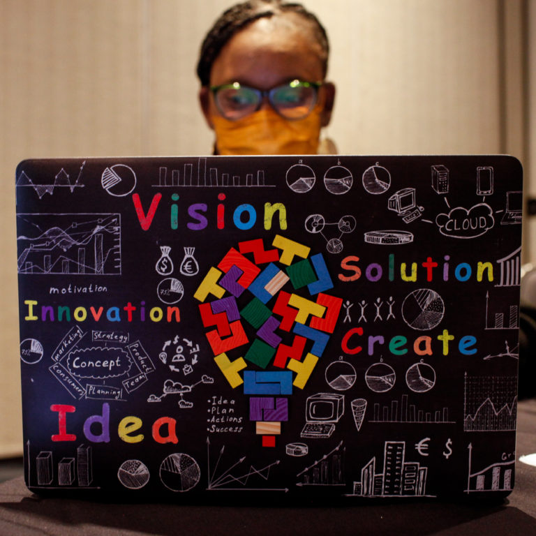 A woman wearing glasses and an orange cloth mask seated at a laptop displayed in the foreground of the image. The laptop has a colorful decorative cover featuring multiple shapes forming a larger lightbulb surrounded by the words: vision, solution, innovation, create, idea