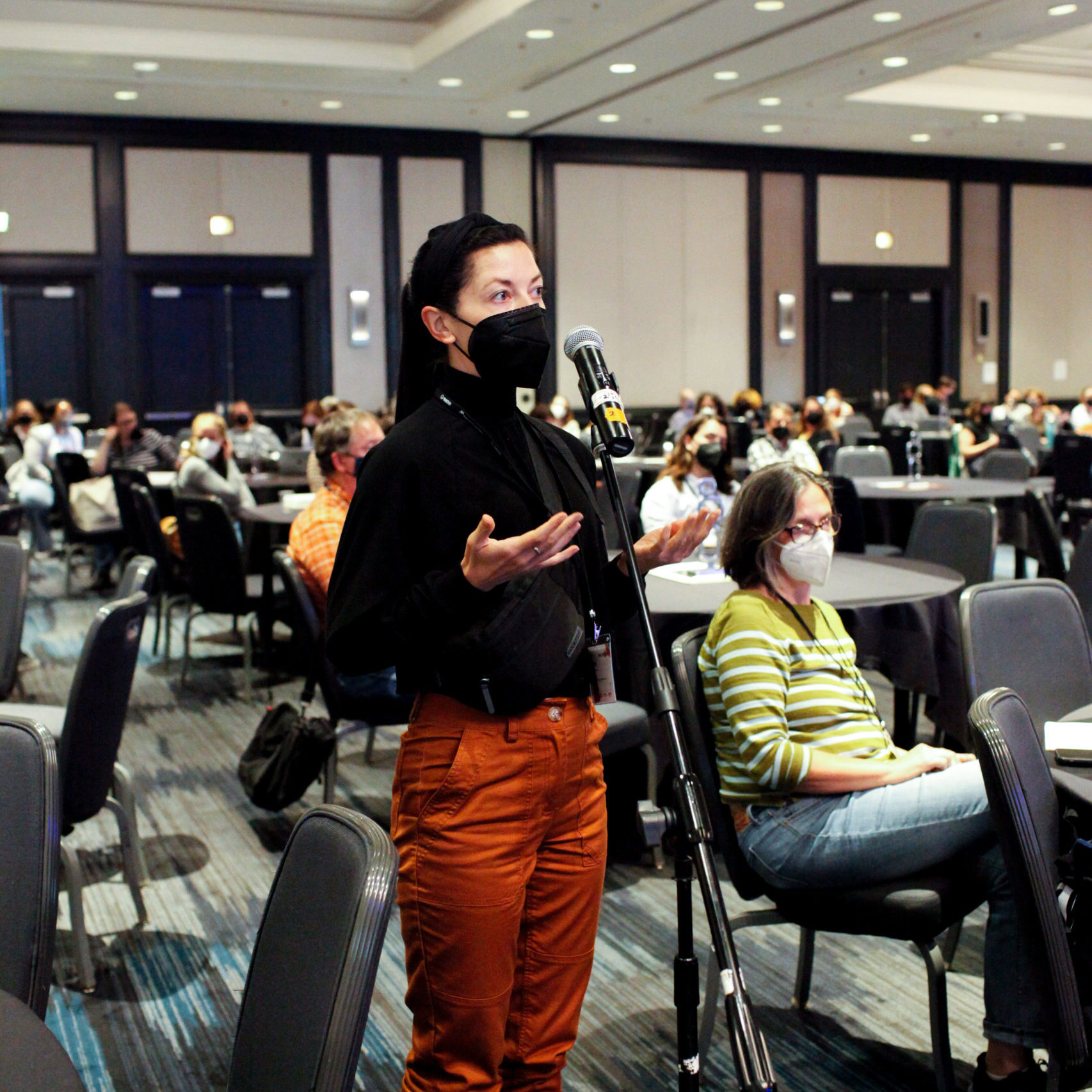 Person wearing an N95 mask at standing at a microphone on a stand. People are seated in a large room in the background.
